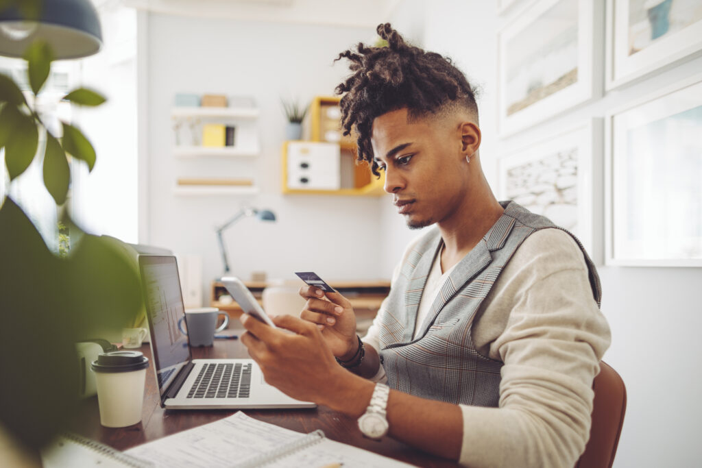 African American man looking at phone and credit card with a laptop sitting in front of him.