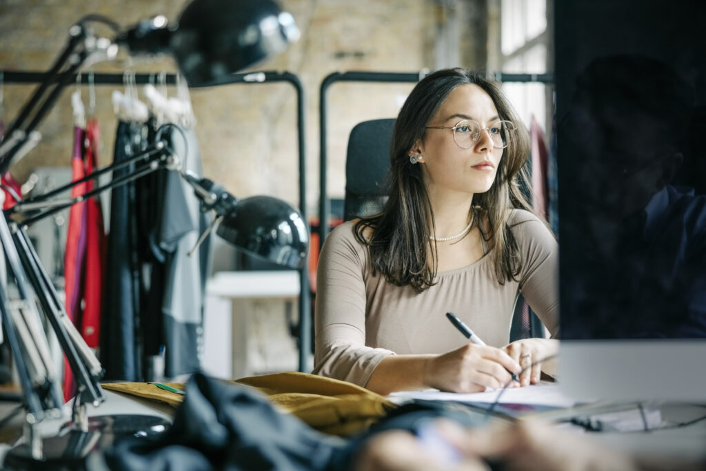 ashion store owner looking at computer screen and writing in a book while working at studio. Fashion designer working at her office.